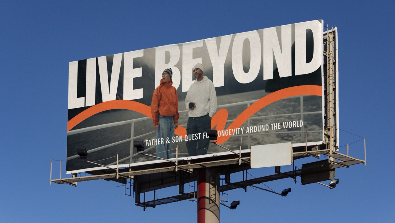 A Live Beyond billboard against a clear blue sky — Marek and Aleksander, and the line: father & son quest for longevity around the world.