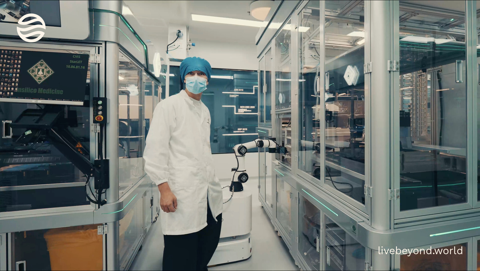 Alek in a white lab coat, cap and mask, standing beside a robot arm among glass-walled automated lab machines.