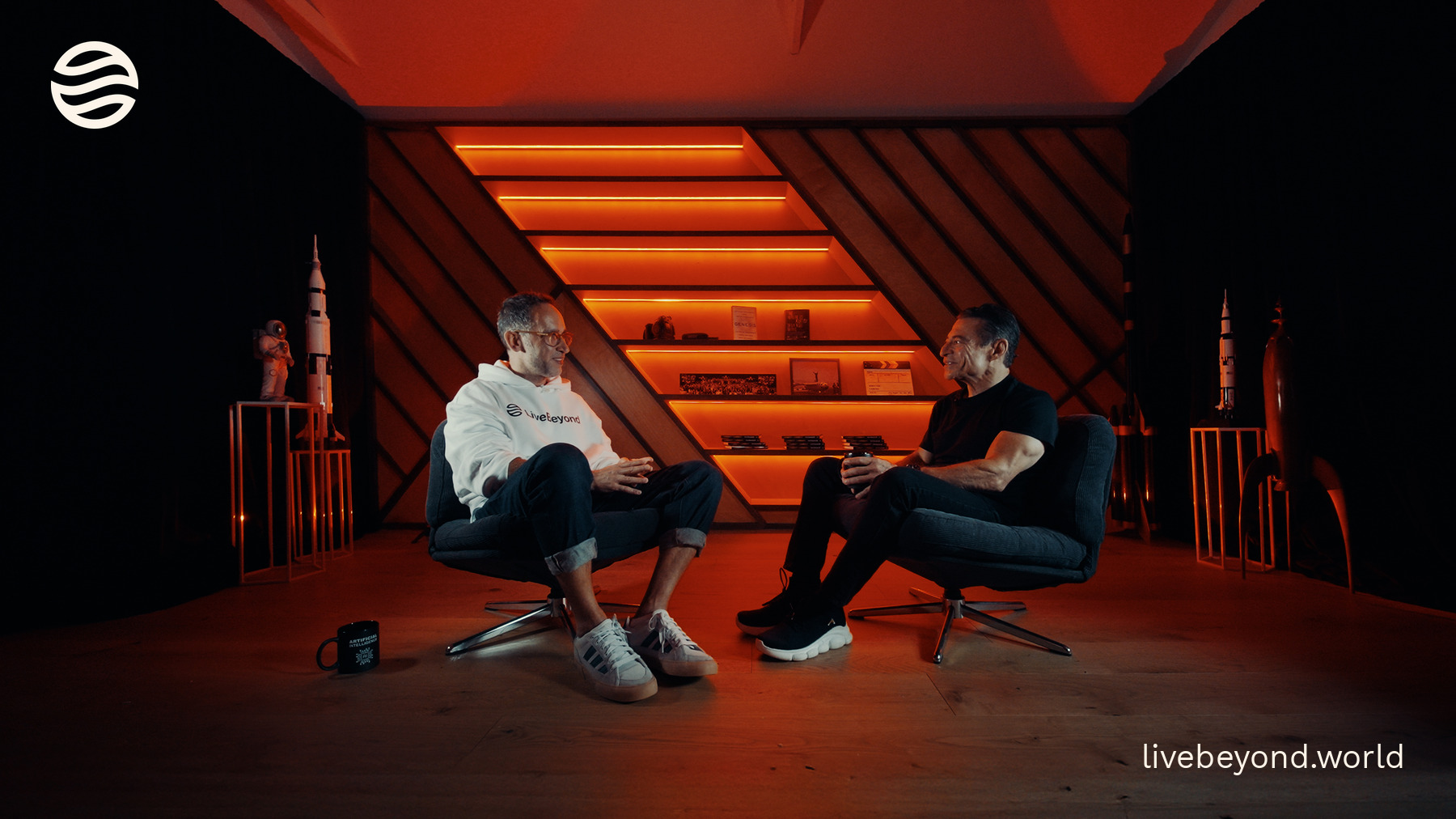 Marek and Peter Diamandis talking in armchairs in a studio glowing orange, with rocket models on the shelves behind them.
