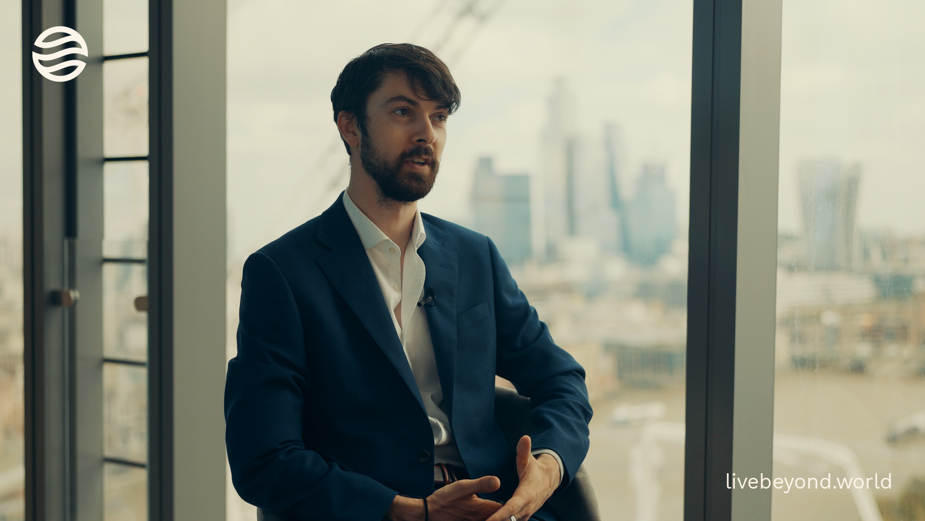 Oliver Zolman in a navy jacket, talking by a tall window with the London skyline behind him.