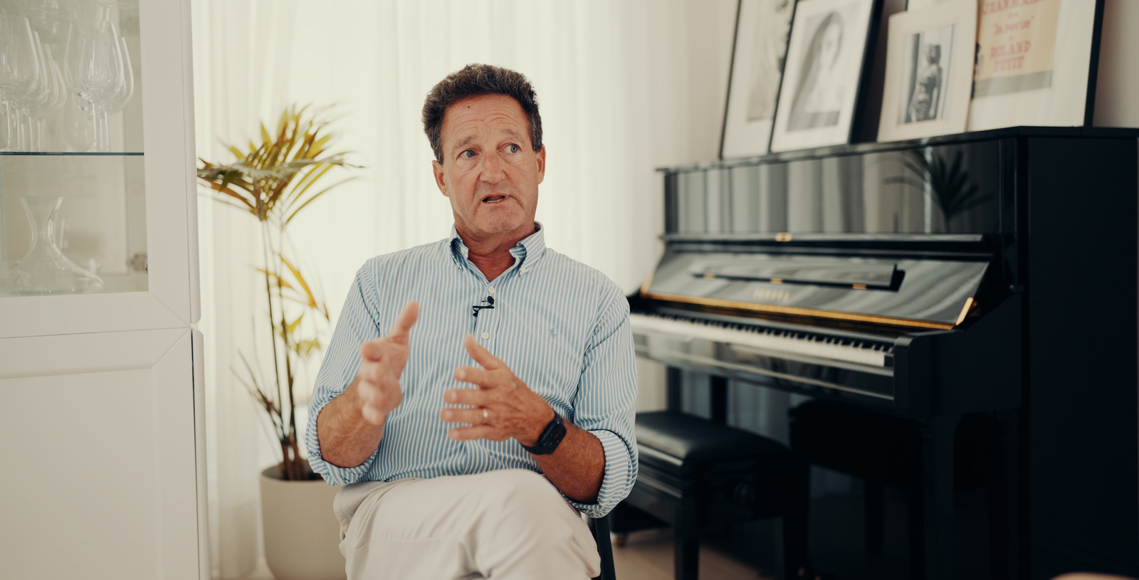 Dr Patrick Sewell explaining with both hands, sitting by a black piano in a bright room in London.