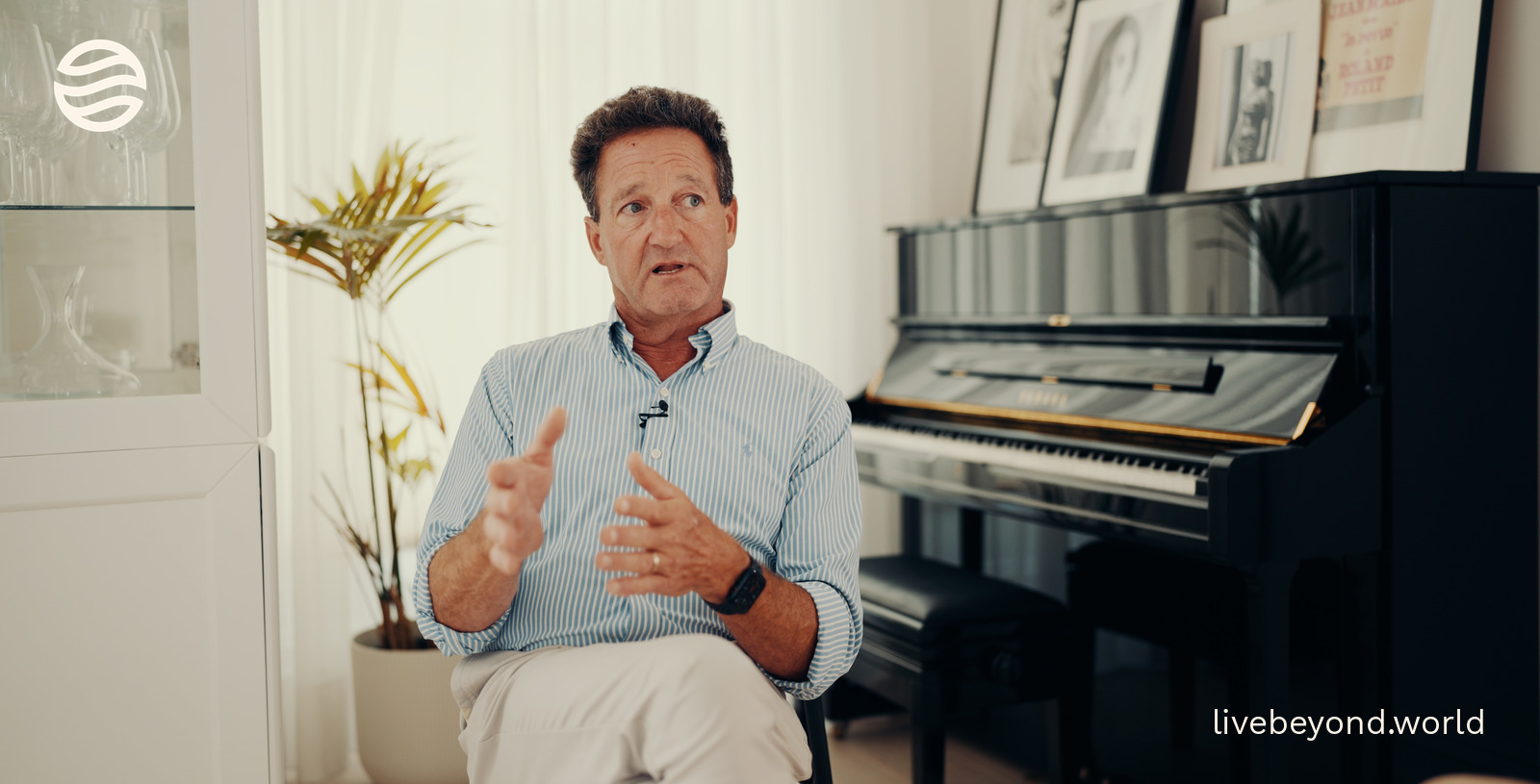 Dr Patrick Sewell explaining with both hands, sitting by a black piano in a bright room in London.