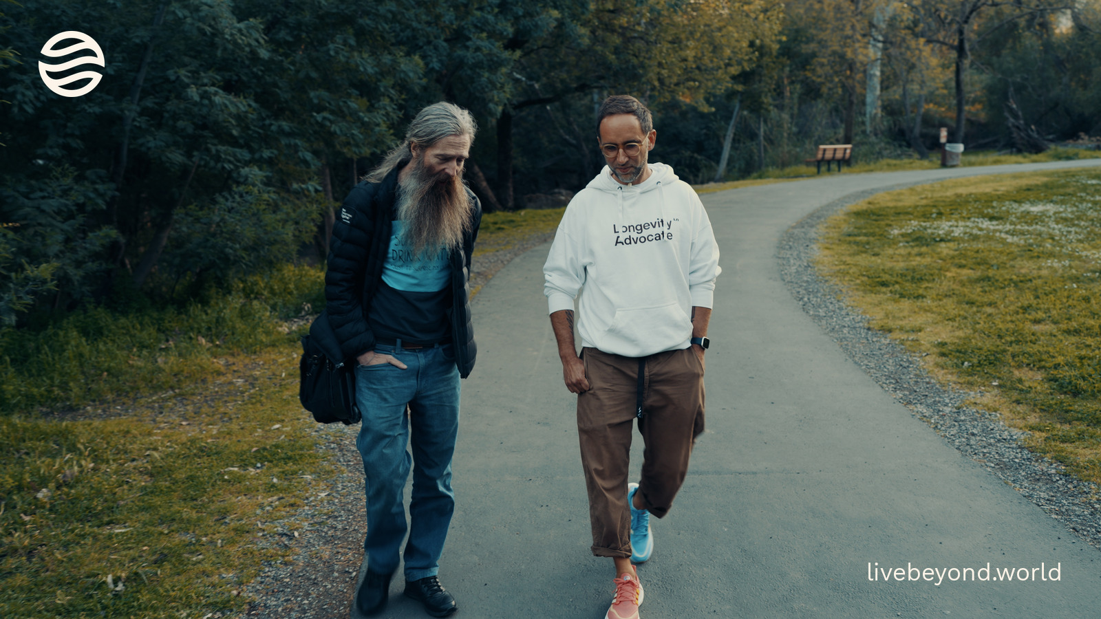 Aubrey de Grey and Marek walking side by side on a curving path through a green park.