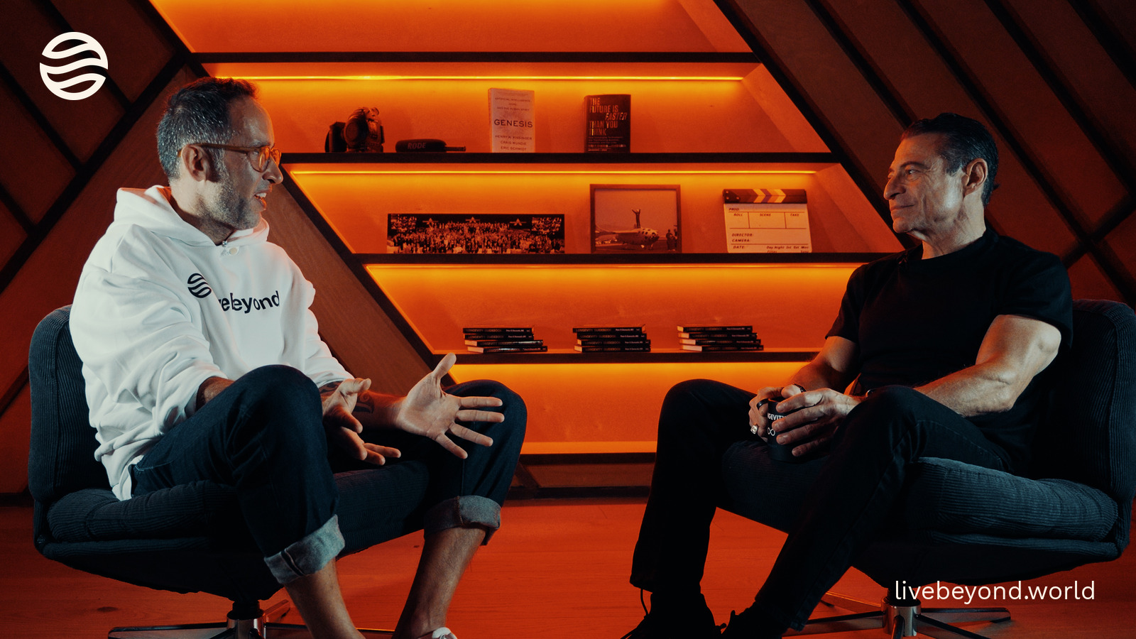 Marek and Peter Diamandis talking in two armchairs in his studio, warm orange shelves behind them.