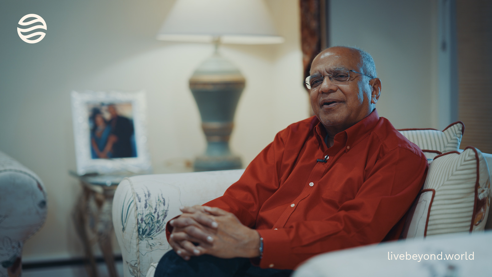 Srikumar Rao in a red shirt, smiling on a sofa in his living room.