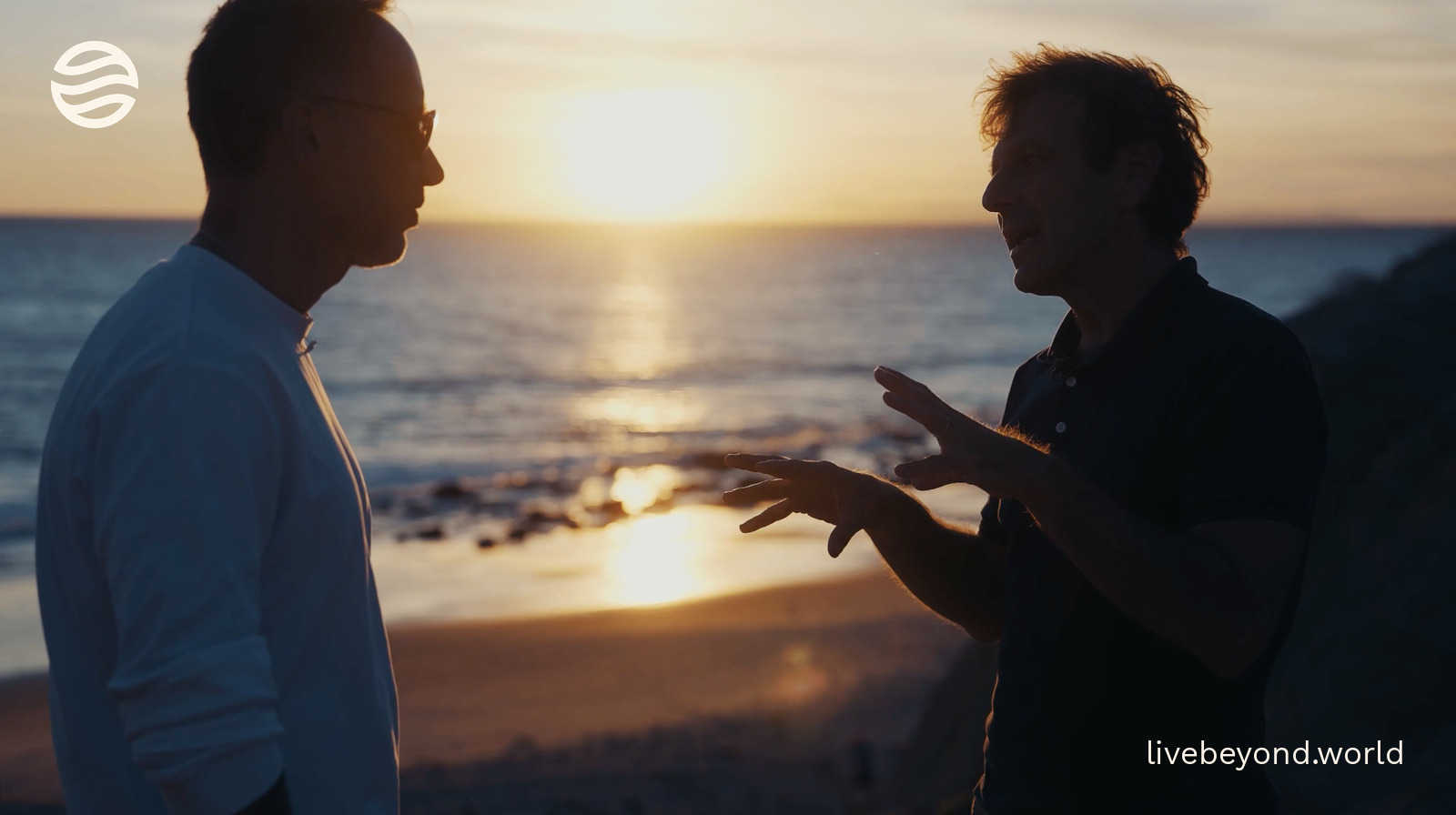 Marek and Vittorio Sebastiano in silhouette, talking on a beach at sunset.