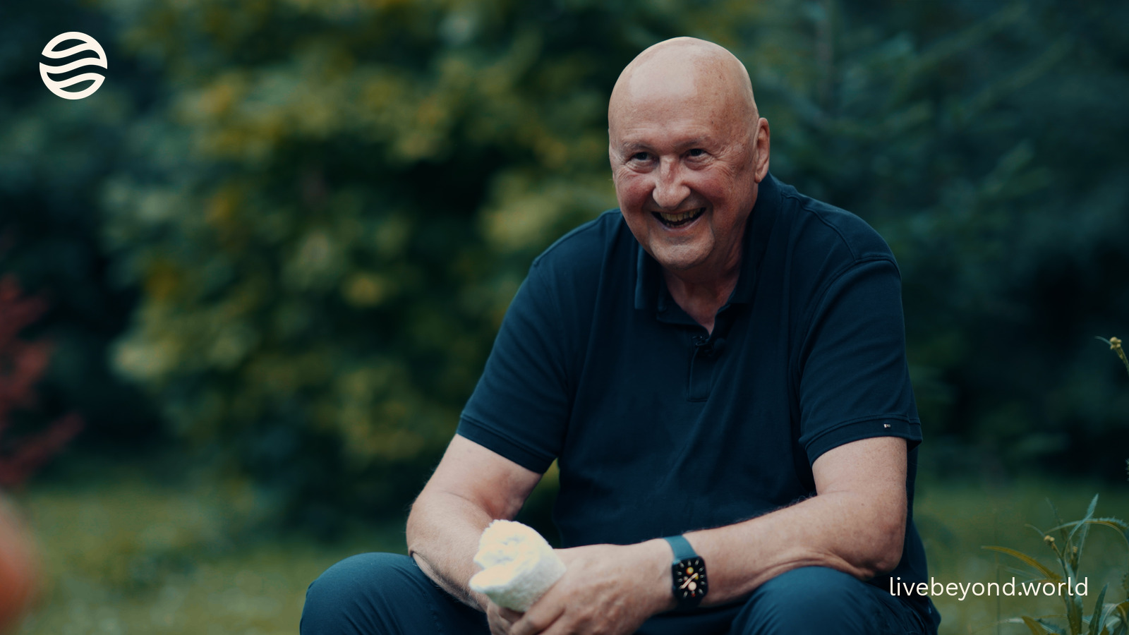 John Davies smiling, sitting in a green wood and holding a small white model.