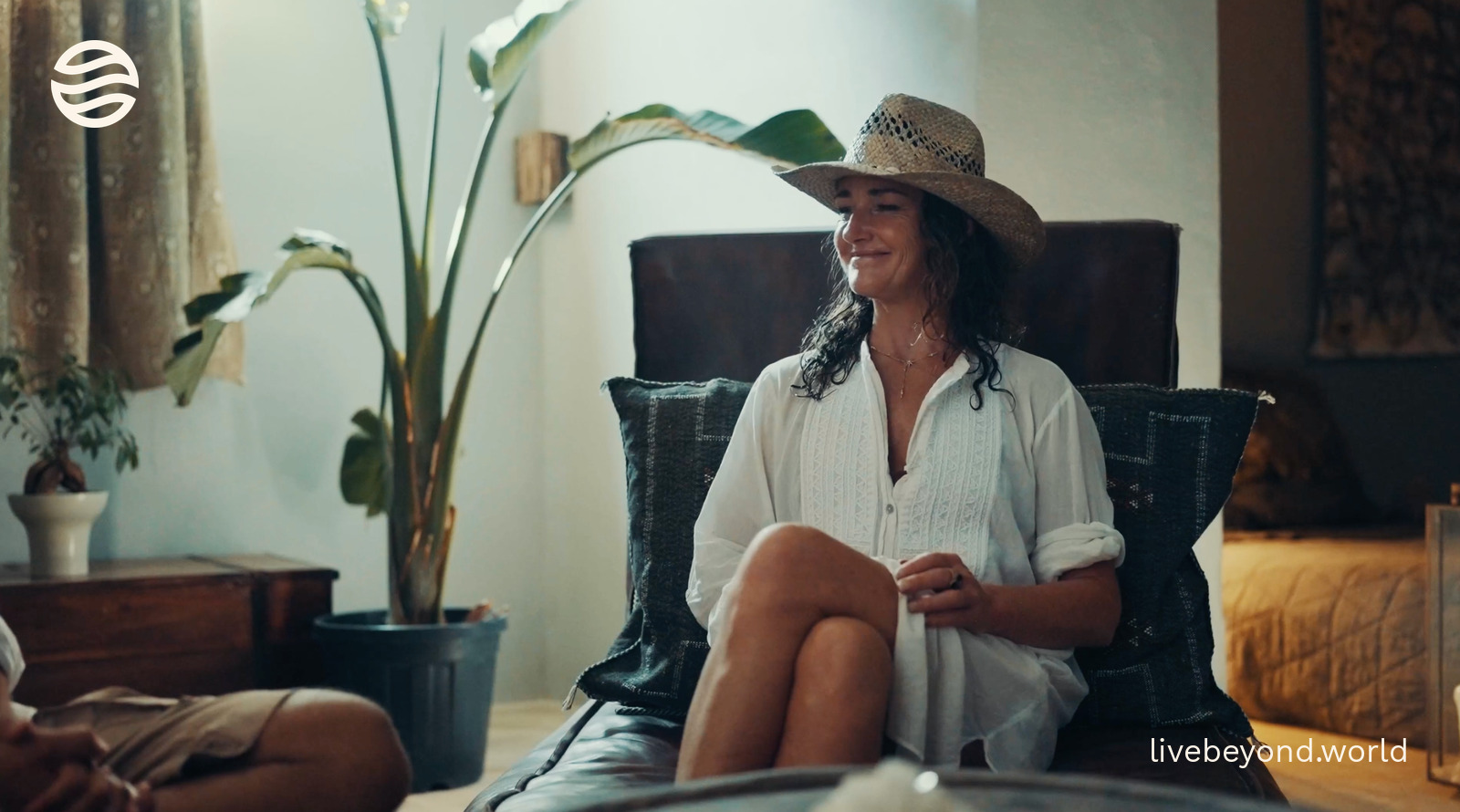 Dr Tamsin Lewis in a straw hat, smiling in an armchair in a bright room with plants.