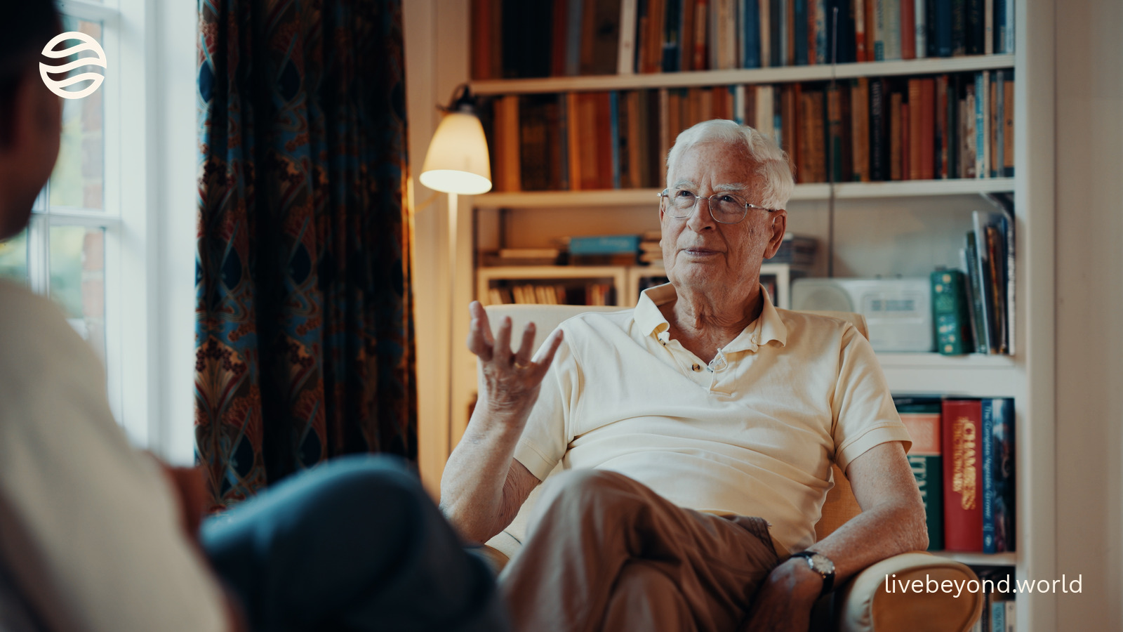 Sir Christopher Ball talking in an armchair in front of bookshelves.