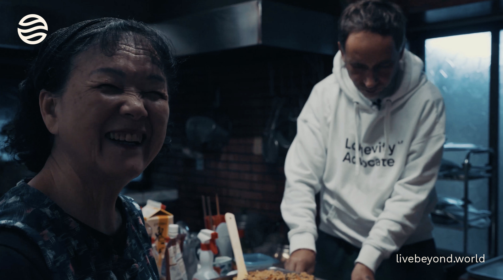 Emi laughing in her café kitchen, with Marek cooking behind her.