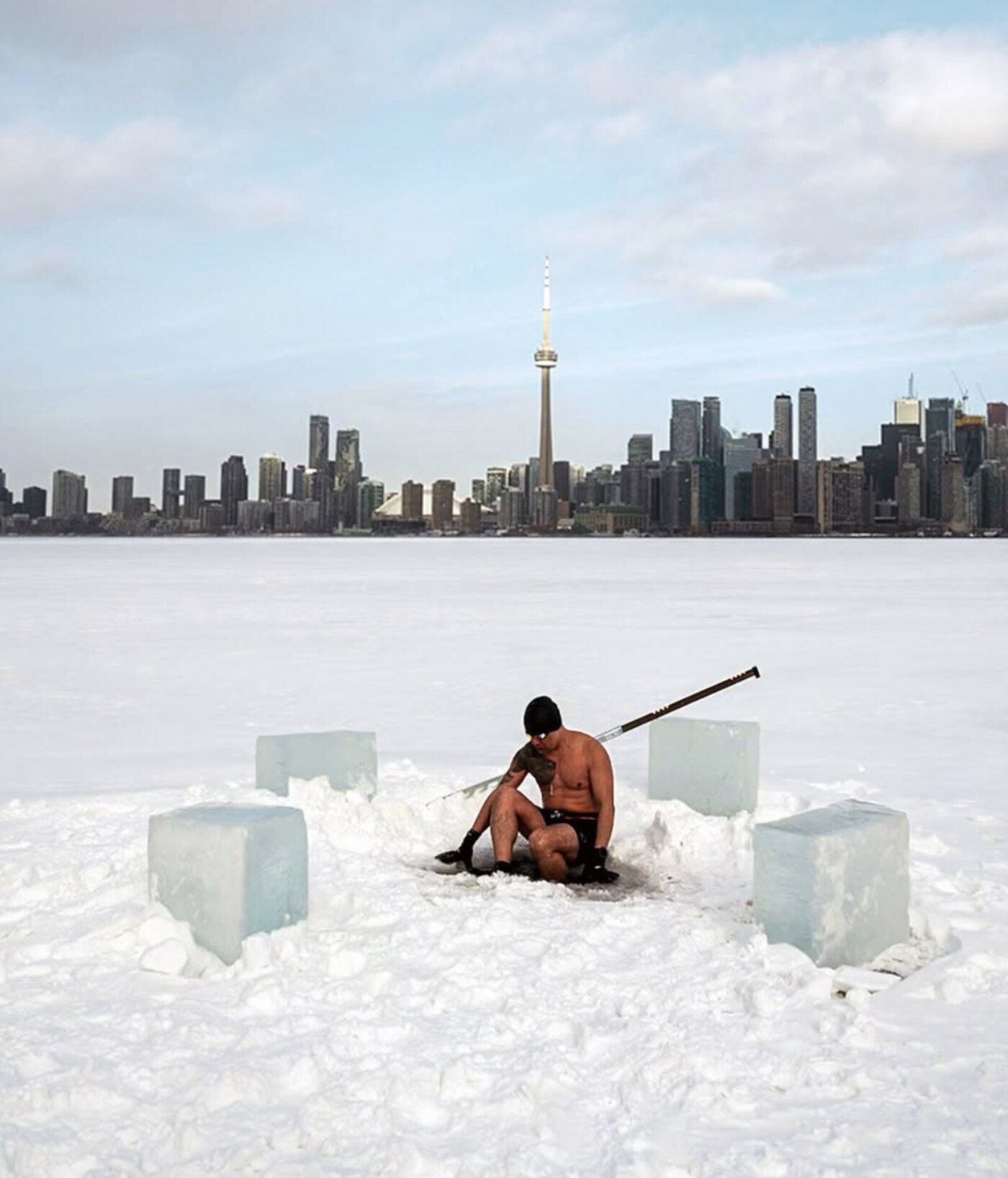 Ron Batuigas sitting in an ice hole he cut, the Toronto skyline behind him